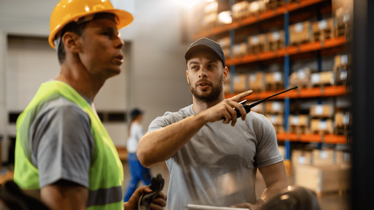 Dos operarios de almacén en conversación activa con equipamiento de seguridad en un entorno de almacén