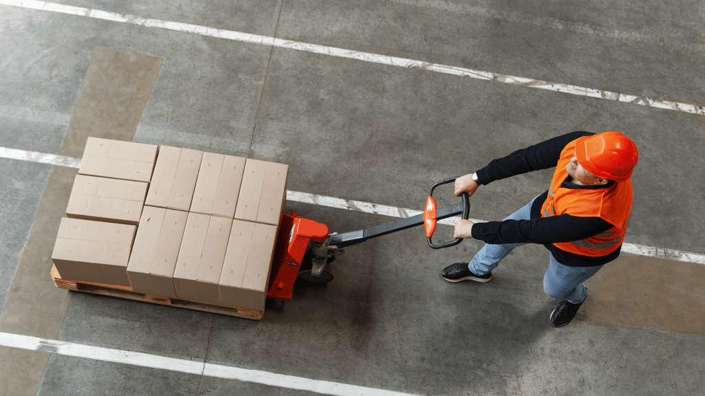 Trabajador con casco de seguridad operando un pallet jack manual en un almacén.